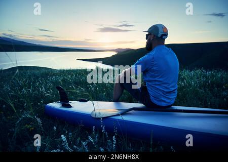 Portrait of bearded Man in silhouette holding SUP board paddle near lake at sunset in Kazakhstan. Stand up paddle boarding outdoor active recreation i Stock Photo
