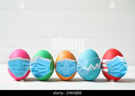 COVID-19 pandemic. Colorful Easter eggs in protective masks on white wooden background Stock Photo