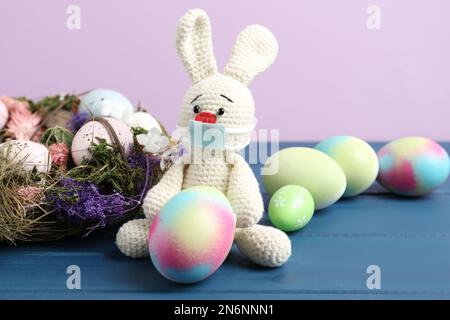 Toy bunny with protective mask and painted eggs on blue wooden table. Easter holiday during COVID-19 quarantine Stock Photo