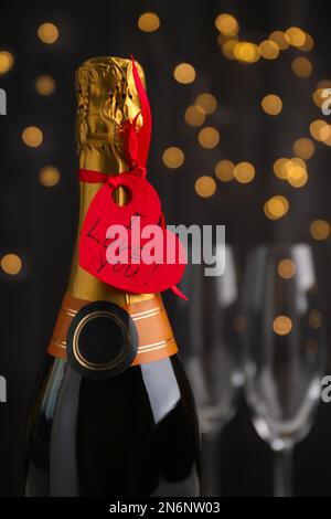 Heart shaped greeting card with phrase I Love You tied to bottle of champagne against blurred lights, closeup Stock Photo