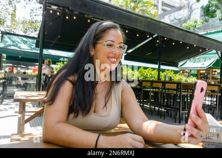 young adult latin woman of venezuelan ethnicity, with glasses and long hair, sitting waiting for her food in the restaurant, smiling using the phone, Stock Photo