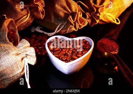 Hessian sacks full of roasted coffee beans with a heart shaped dish full of beans and a coffee scoop full of ground coffee Stock Photo