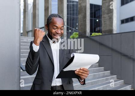 Happy african american businessman, lawyer, banker standing near office and holding folder with papers. He reads documents, shouts with joy, shows a victorious yes gesture. Stock Photo