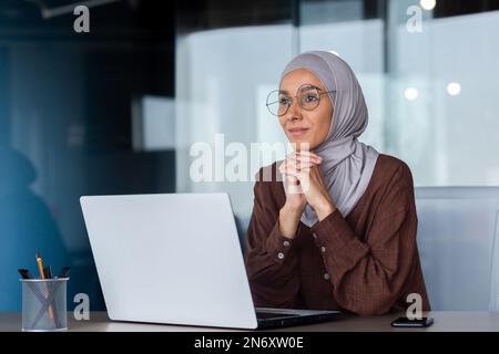 Portrait of a young Arab woman in a hijab and glasses sitting in the office at a table, working on a laptop. She rested her head on her hands. Thoughtfully and dreamily looks ahead, out the window. Stock Photo