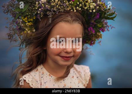 Cute little girl wearing wreath made of beautiful flowers on blurred background, closeup Stock Photo