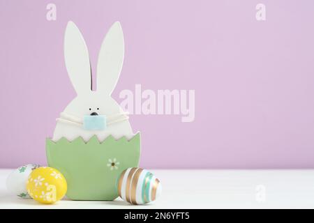 Wooden bunny with protective mask, painted eggs and space for text on white wooden table. Easter holiday during COVID-19 quarantine Stock Photo