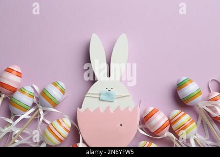 Wooden bunny with protective mask and eggs on lilac background, flat lay. Easter holiday during COVID-19 quarantine Stock Photo