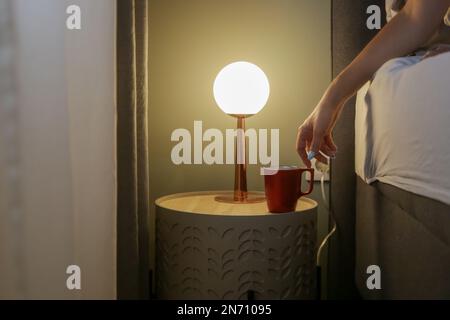 Woman in bed reaching for her first morning coffee from the nightstand. A red mug on the bedside table Stock Photo
