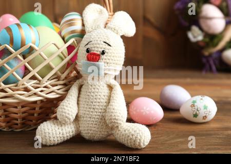 Toy bunny in protective mask near dyed eggs on wooden table. Easter holiday during COVID-19 quarantine Stock Photo