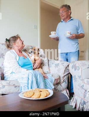 Retirement: Tea and Biscuits. A senior man bringing his wife a cup of tea as she sits with their pet dog on the sofa. From a series of related images. Stock Photo