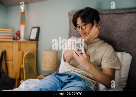 Young asian man in earphones using mobile phone while resting on bed at home Stock Photo