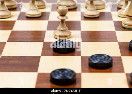 playing by different rules on the same board - black checkers and white chess figures on wooden chessboard, pawn and checkers piece in center of playf Stock Photo