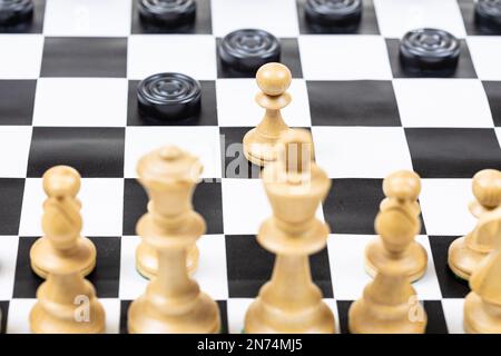 playing by different rules on the same board - black checkers and white chess figures on black white chessboard, pawn and checkers disc moves closeup Stock Photo