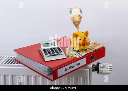 Red file folder with label energy costs, pocket calculator, piggy bank, various euro bills lying on radiator, 1350 euros, symbol image, heating bill, energy costs, rising heating costs, white background, Stock Photo