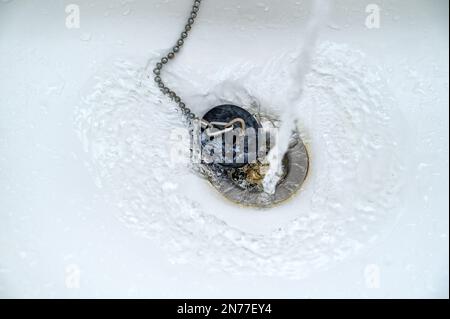 Water running in a white wash basin or bathtub with an old black rubber plug and a rusty drain, copy space, selected focus, narrow depth of field Stock Photo