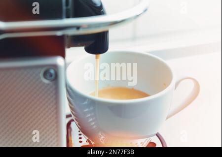Fresh hot coffee runs out of a modern capsule machine into a white porcelain cup, colored light reflexes, bright background with copy space, selected Stock Photo