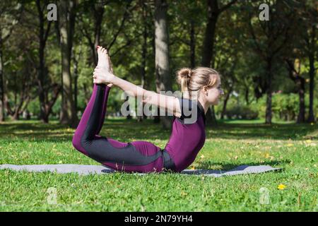 attractive slender white blond girl in violet overall practice iyengar yoga complex in summer sunny green park and demonstrates bow pose dhanurasana Stock Photo