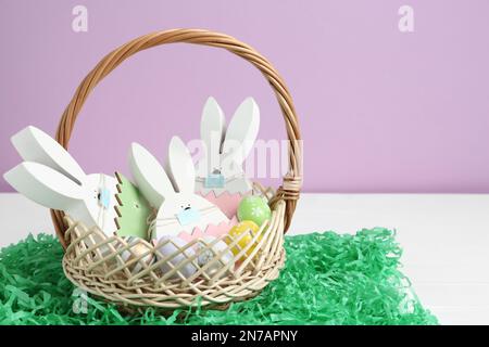 Wooden bunnies with protective masks, painted eggs in basket and paper grass on white wooden table. Easter holiday during COVID-19 quarantine Stock Photo
