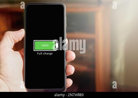 Close up of male hand holding smartphone with full battery icon on the screen indicating that battery level is at one hundred percent. Full charged ba Stock Photo