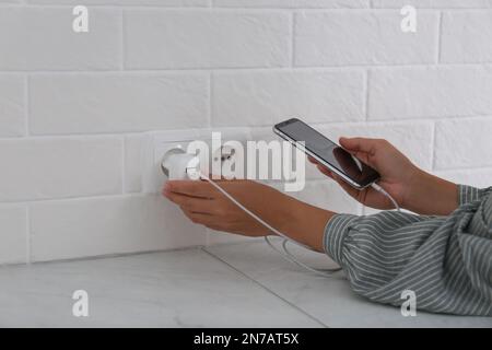 Woman with mobile phone plugging charger in socket on white brick wall, closeup Stock Photo