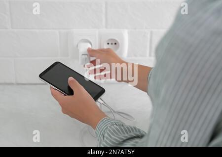 Woman with mobile phone plugging charger in socket on white brick wall, closeup Stock Photo