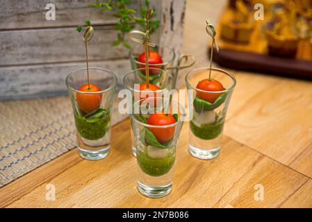 Caprese canapes in glass stacks. Cherry tomatoes, mini mozzarella balls, pesto sauce and basil leaves in a glass on a wooden table. Stock Photo