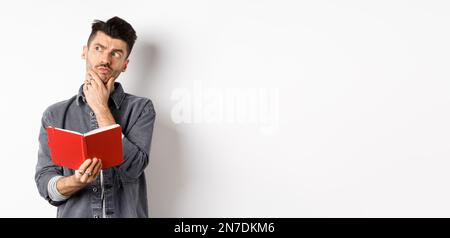 Thoughtful young man plan his schedule, looking aside pensive, holding opened planner or journal, standing on white background Stock Photo