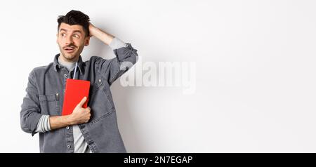 Confused guy scratch head and look aside at logo puzzled, holding red diary or planner, standing against white background Stock Photo