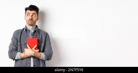 Silly young man looking aside at logo with timid face, holding his diary pressed to chest, carry journal, standing on white background Stock Photo