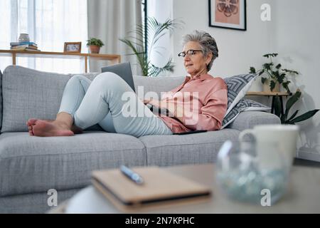Senior woman, laptop and relax on sofa in the living room checking email, typing or writing at home. Elderly female freelancer or writer relaxing on Stock Photo