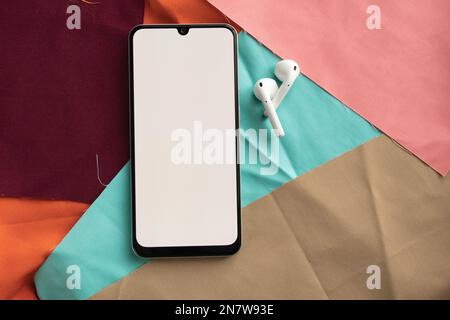 a phone with a white screen and wireless headphones lies on lies on multi-colored fabrics close-up Stock Photo