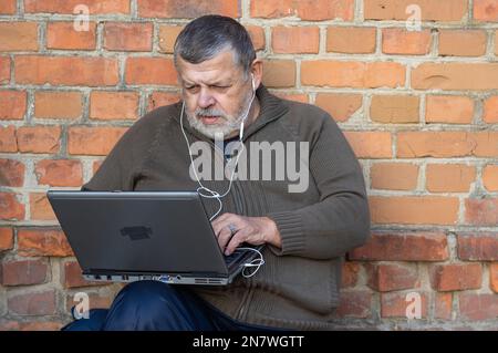 Nice portrait of bearded senior man working on a laptop and listening music while sitting against brick wall Stock Photo