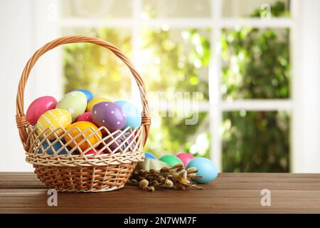 Colorful Easter eggs in wicker basket and willow branches on wooden table, space for text Stock Photo