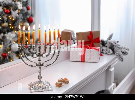 Silver menorah near dreidels with symbols He, Pe, Nun, Gimel on white mantelpiece in room decorated for Christmas Stock Photo