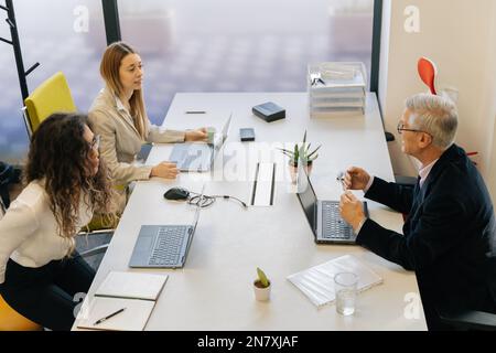 Two junior female coworkers are struggling with the task. Their senior boss is helping them Stock Photo