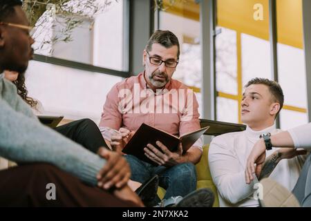 MIddle-aged man reading some notes he took about the project. Tired junior worker is listening to him Stock Photo