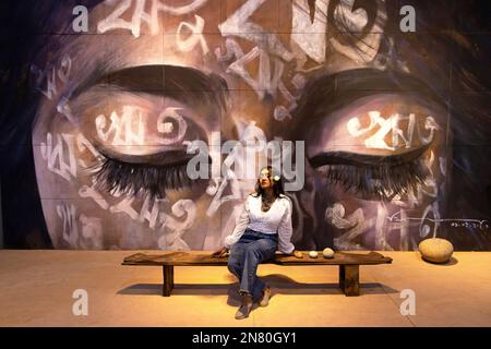 Dhaka, Dhaka, Bangladesh. 11th Feb, 2023. A woman poses in front of an artwork - ''Reetu'' - by Bishwajit Goswami during Dhaka Art Summit at National Art Gallery in Dhaka, Bangladesh. Written in Bengali across the face, the word - ''Reetu'', meaning ''˜seasons', is also a commonly used first name for Bengali girls, culturally and symbolically related to the name ''˜Bonna' (Flood) which is the theme of Dhaka Art Summit. Bangladesh has six seasons (and some would argue 'had'' as climate change has made two seasons difficult to recognize anymore) each harkening to a particular mood, fee Stock Photo