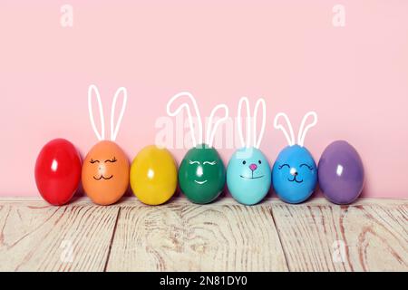 Several eggs with drawn faces and ears as Easter bunnies among others on white wooden table against pink background Stock Photo