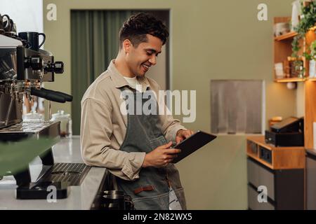 Side view of a smiling barista in an apron typing on a digital tablet leaning counter in a coffee shop Stock Photo