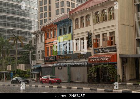 A row of colorful historic colonial mansions with shops and restaurant signs and cars driving down the street along them on a rainy day in Singapore. Stock Photo