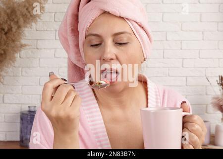 funny woman in a bathrobe after a shower eats a delicious dessert for breakfast. middle-aged woman is in the kitchen enjoying her morning meal. Stock Photo