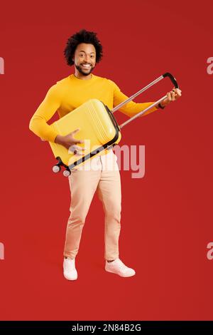 Overjoyed 30s guy holding yellow suitcase and pretend playing guitar, ready for adventure and holiday, dancing and fulling around isolated on red background, travel and vacation concept. Full length Stock Photo