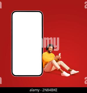Curly African-American curly guy sit near big huge smartphone with blank screen, copy space, mockup, full length young handsome man in yellow pullover using mobile phone, texting near big phone Stock Photo