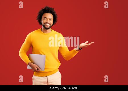Cheerful handsome curly african-american guy hold laptop and point with palm aside at empty space isolated on plain red background, man in yellow advertising, recommending new computer app Stock Photo