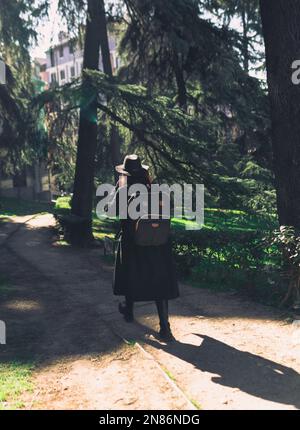 A vertical back view of a young female wearing black coat and backpack in a park Stock Photo