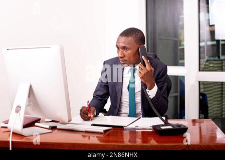 businessman sitting in front of computer and talking on mobile phone while taking notes in document in office. Stock Photo
