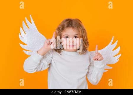 Child angel pointing up, point gesture, idea. Valentine's day. Blonde cute child with angel wings on a yellow studio background. Happy angel child. Stock Photo