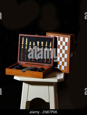 Small wooden chess set alongside a black flask on a white table with black background. Stock Photo