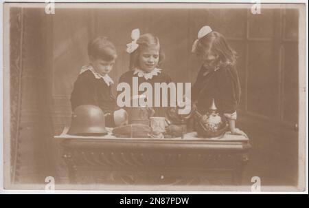 Three British young children looking at First World War trophies taken from German soldiers. The objects laid out on the table include a German Pickelhaube helmet, a Stahlhelm helmet, a field cap, a gas mask and a tin cup or mug. The photograph was taken by the Tasma Studios, 99 Wellington Street, Woolwich, London Stock Photo