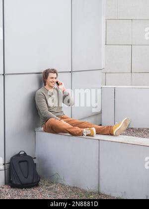 Smiling man sits talks by smartphone on outdoor part of wall. Student with rucksack in casual clothes. Handsome man with long hair. Stock Photo